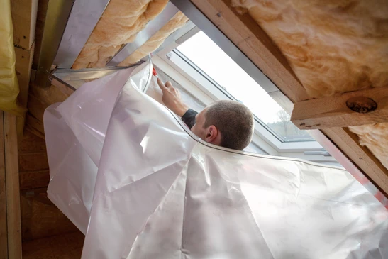 Un homme installe un film plastique de protection autour d'un châssis de fenêtre de toit Velux dans des combles en construction