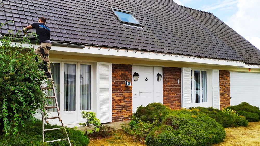 Artisan en tenue de travail bleue, debout sur une échelle aluminium, intervenant sur une gouttière en bordure de toiture