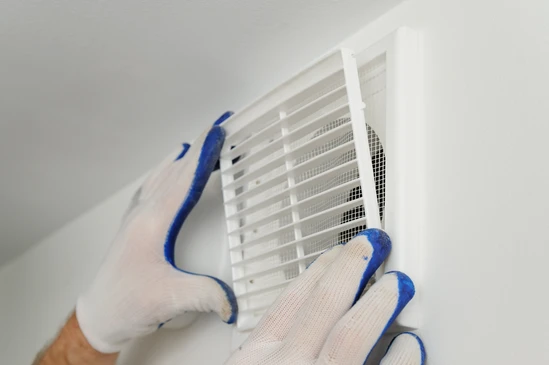 Main gant&eacute;e tenant une grille d'a&eacute;ration blanche avec lamelles orientables et moustiquaire int&eacute;gr&eacute;e, fixation murale ou plafond visible