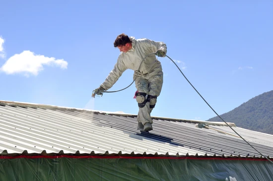 Ouvrier en combinaison de protection appliquant un revêtement sur toiture métallique ondulée à l'aide d'un pistolet à peinture relié par flexible