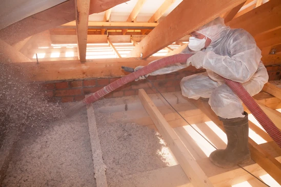 Professionnel en combinaison de protection intégrale avec masque respiratoire travaillant dans combles, aspirant isolant avec tuyau flexible rouge connecté à équipement d'extraction