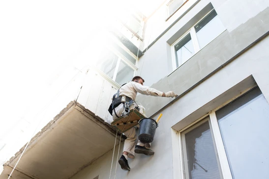 Ouvrier en tenue blanche effectuant des travaux de ravalement sur façade extérieure d'immeuble, suspendu par harnais de sécurité